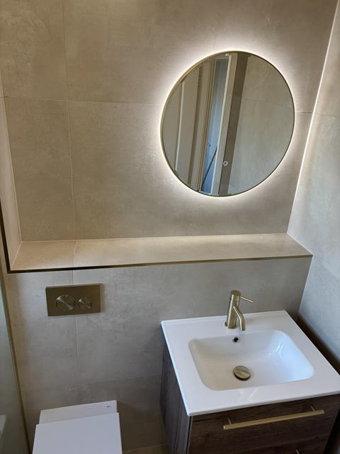 Backlit round mirror, brushed brass tap and flush plate, and stone-effect tiles with a tiled ledge
